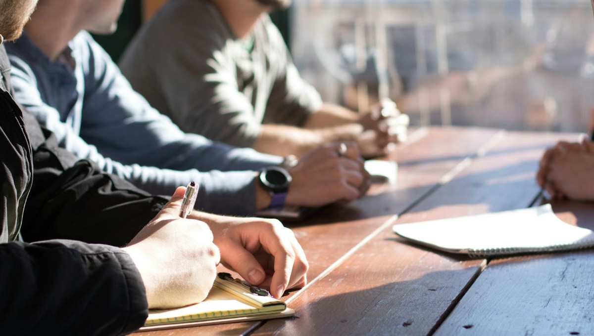 Personer träffas och pratar under ett par träd, solen lyser in mellan träden. Några hundar står och väntar, en person cyklar iväg